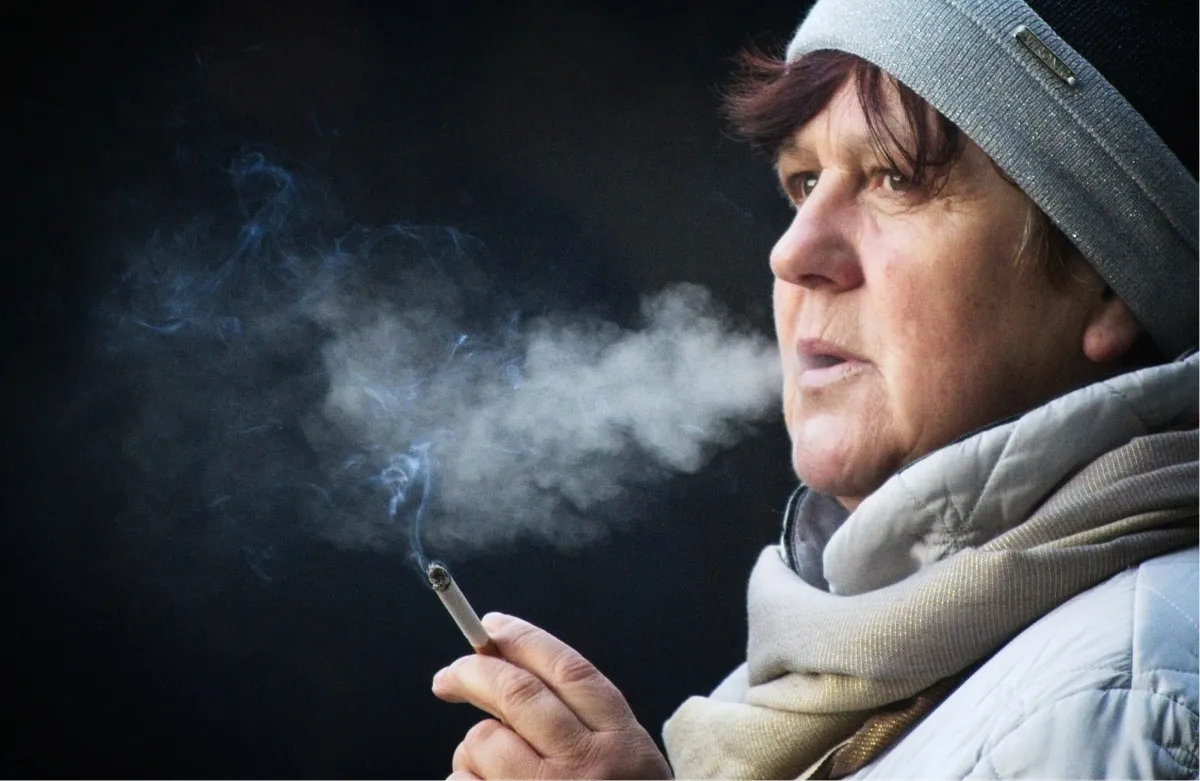 Older woman in a knit hat holding a cigarette as smoke drifts across her face